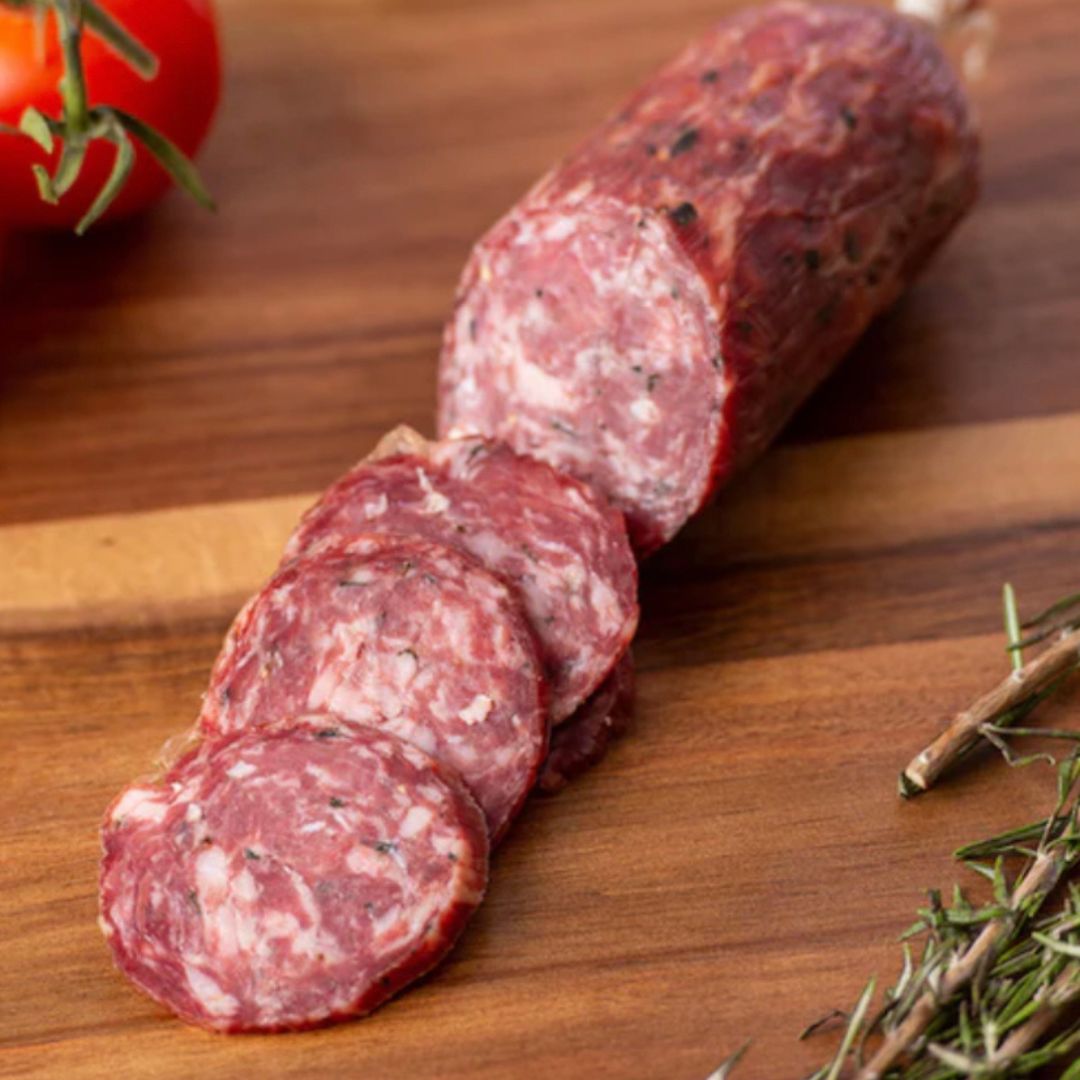 Wild Boar Bush Tucker Salami on a wooden board, a tomato on the vine in the top left corner and rosemary sprigs in the bottom right corner
