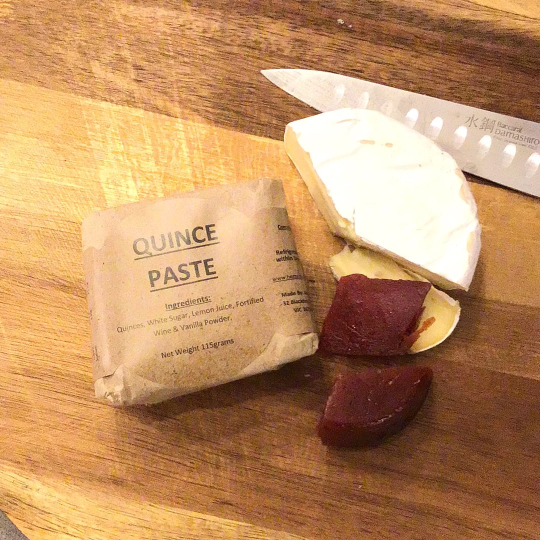Quince paste jar, wrapped in brown paper. Placed on a wooden board alongside Brie cheese and two pieces of quince paste