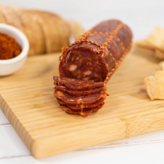 Chorizo thinly sliced on a wooden board with a bowl of paprika, crackers and loaf of bread in the background