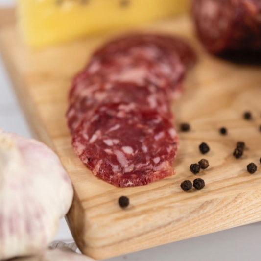 Veneto Cracked Pepper and Garlic Salami on a wooden board surrounded by peppercorns and garlic cloves