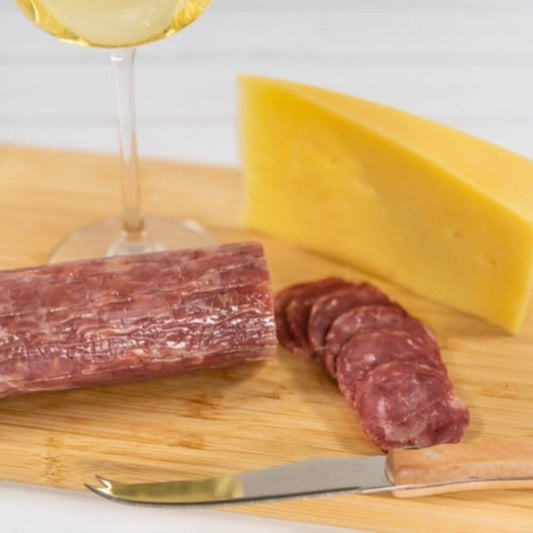Duck and juniper berry salami on a wooden board, cut into thing slices. Cheese knife in the forefront, block of cheese and glass of white wine in the background