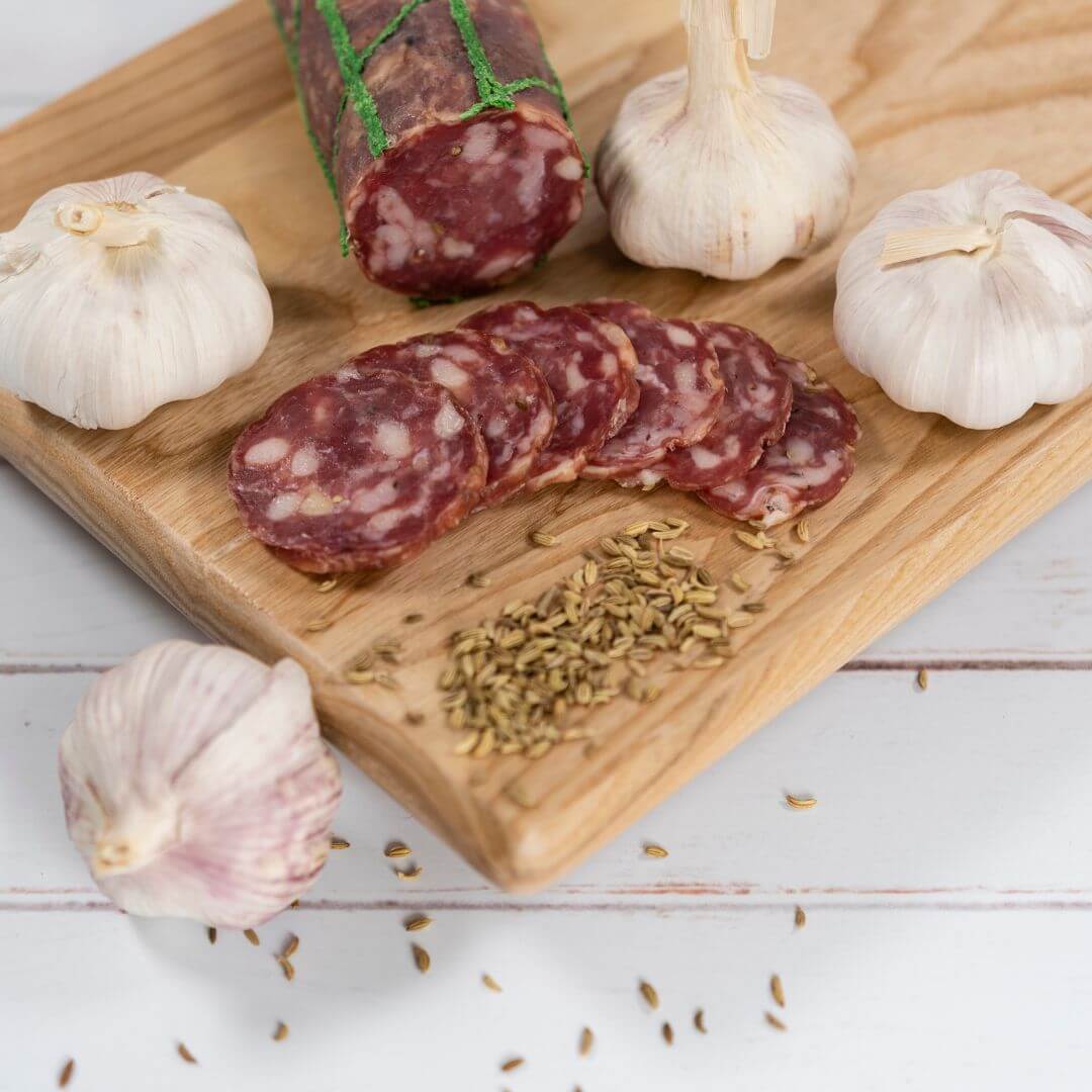 Thinly sliced fennel and garlic pork salami on a wooden board with garlic and fennel seeds surrounding it