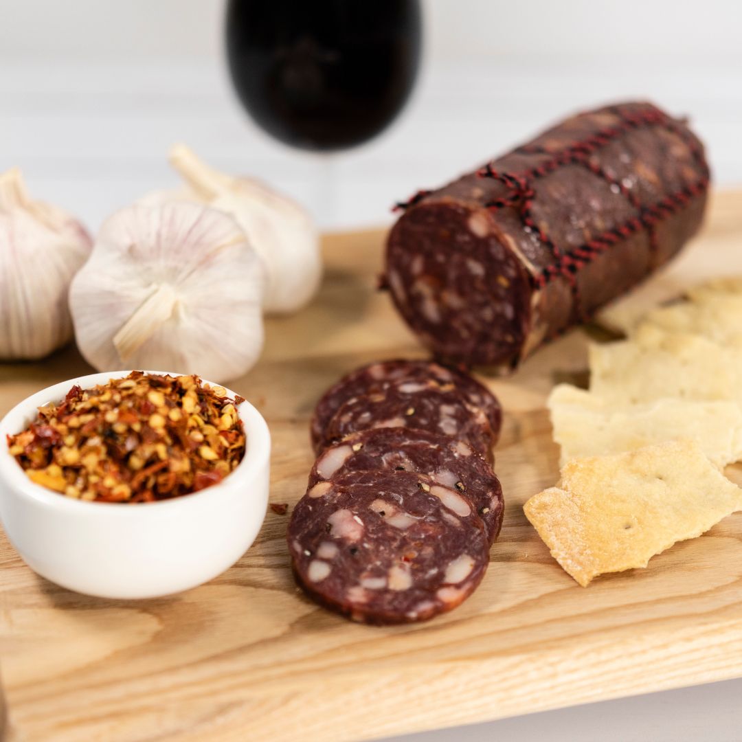 Kangaroo salami thinly sliced on a wooden board with garlic cloves, crackers, a bowl of chilli flakes and a glass of red wine