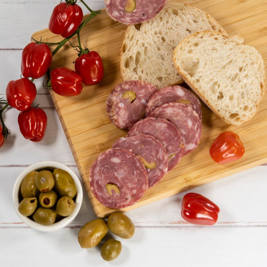 Olive Salami, thinly sliced and placed on a wooden board with two slices of bread, cherry tomatoes on the vine and a small bowl of green stuffed olives